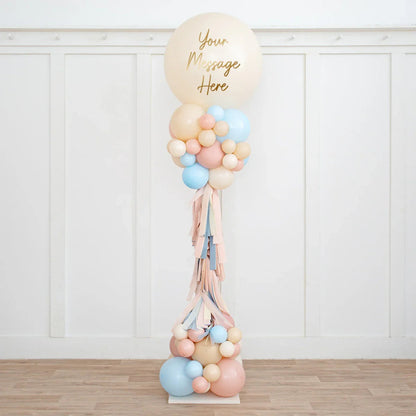 Tall balloon pillar with personalized message in beige, baby pink, and blue colors, indoor wooden floor