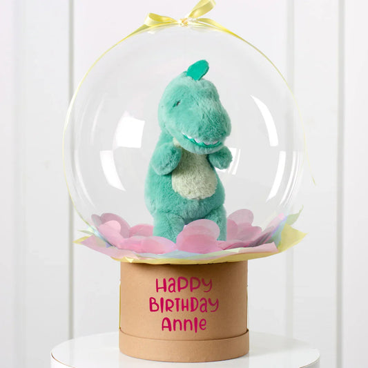 Green plush dinosaur inside clear balloon with pastel confetti on birthday gift box