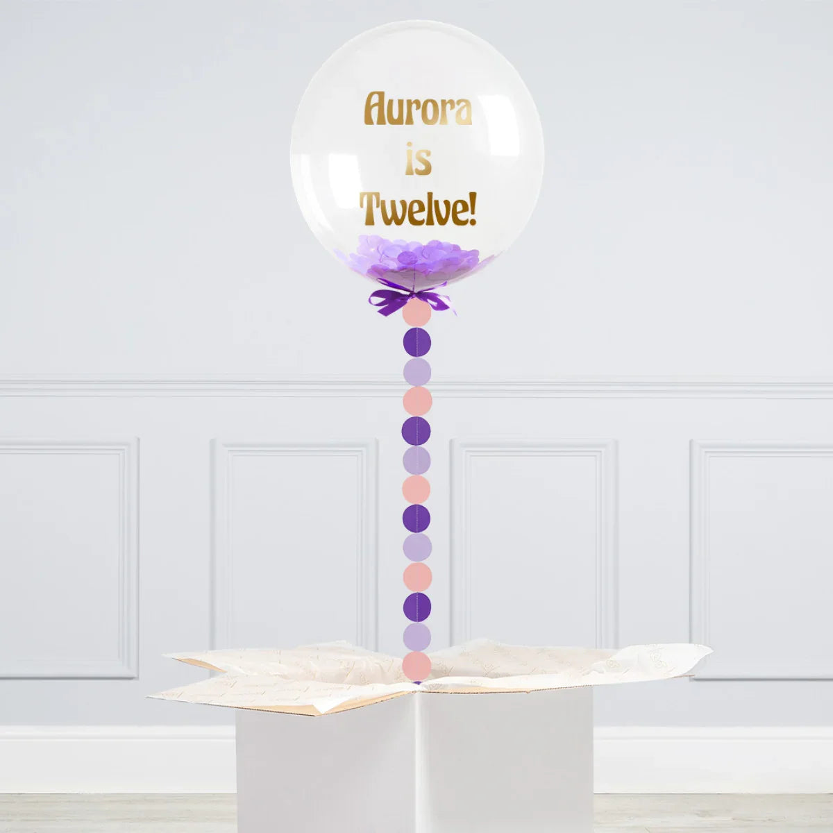 Personalized clear bubble balloon with purple confetti and gold text 'Aurora is Twelve!' attached to a gift box