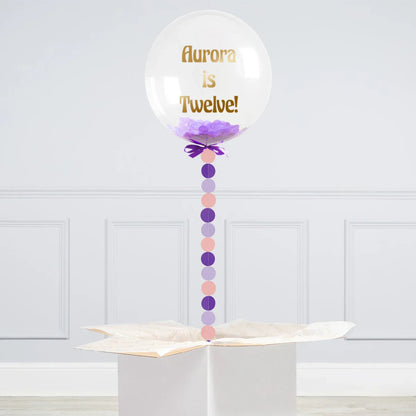 Personalized clear bubble balloon with purple confetti and gold text 'Aurora is Twelve!' attached to a gift box
