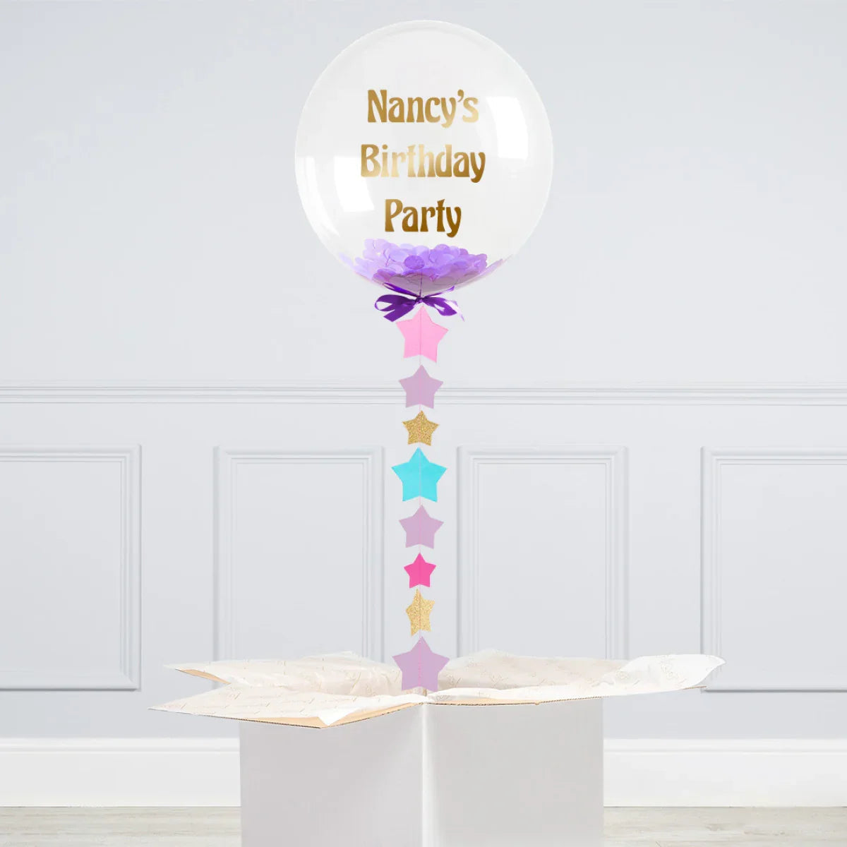 Clear balloon with purple confetti and gold text 'Nancy's Birthday Party' floating above white gift box
