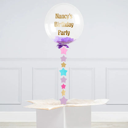 Clear balloon with purple confetti and gold text 'Nancy's Birthday Party' floating above white gift box