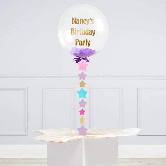 Clear balloon with purple confetti and gold text 'Nancy's Birthday Party' floating above white gift box