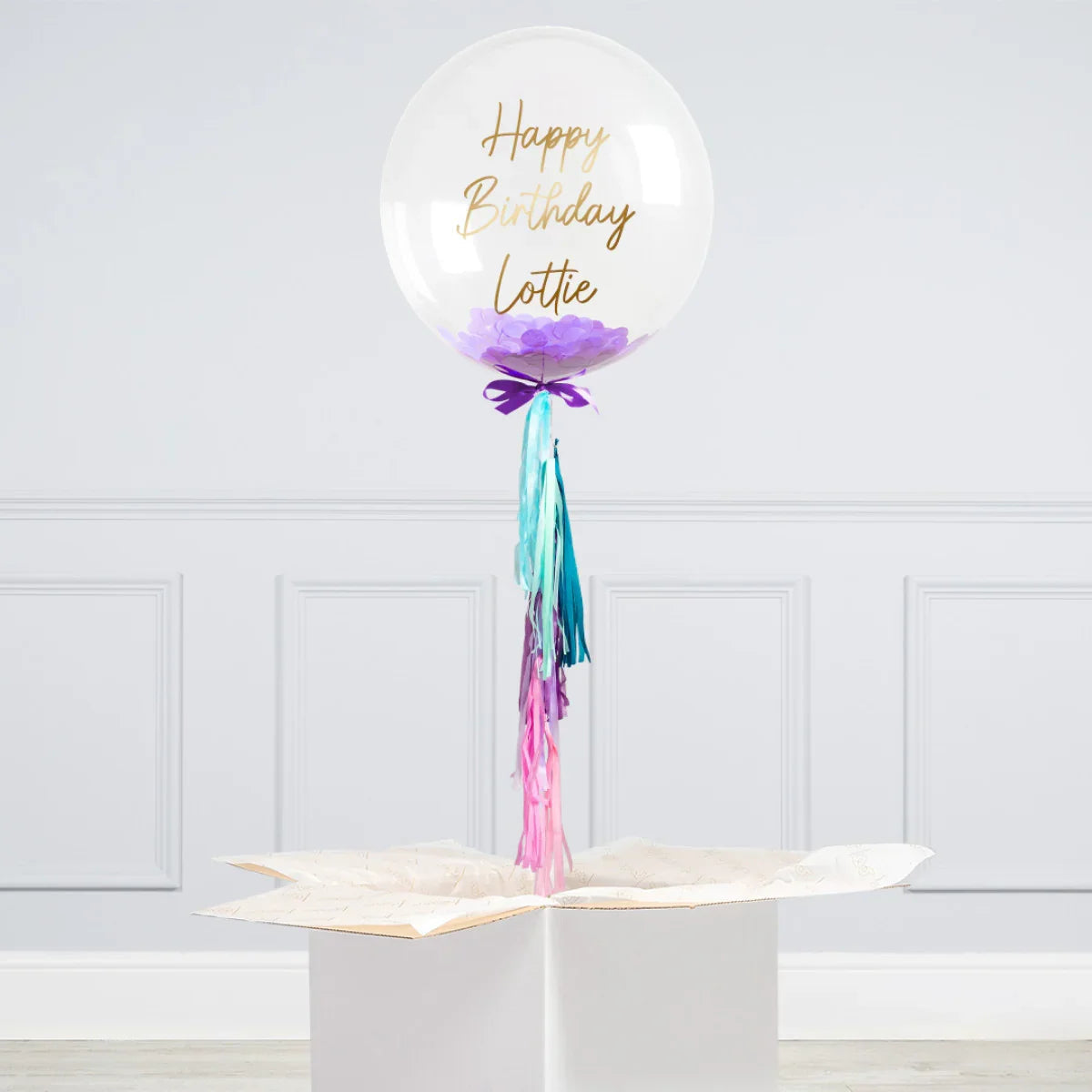 Clear personalised helium bubble balloon with purple confetti and Happy Birthday Lottie text, tied with colorful tassels