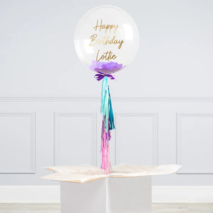 Clear personalised helium bubble balloon with purple confetti and Happy Birthday Lottie text, tied with colorful tassels