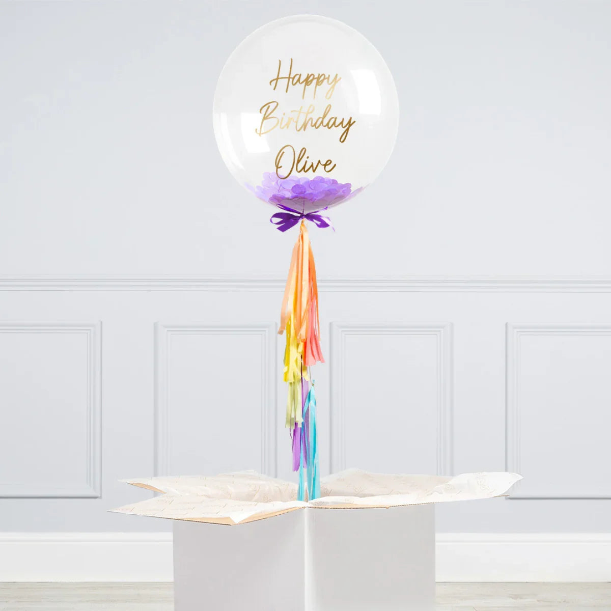 Clear personalized birthday bubble balloon with purple confetti and colorful tassels standing in white gift box