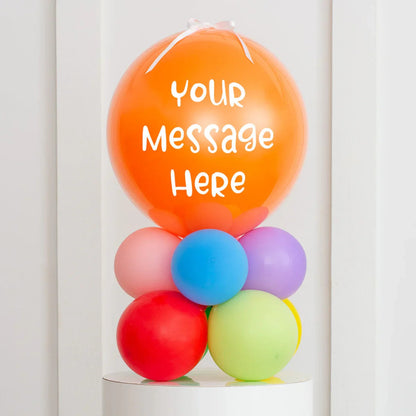 Personalised orange air-filled bubble balloon with white ribbon and multi-coloured smaller balloons base on white pedestal