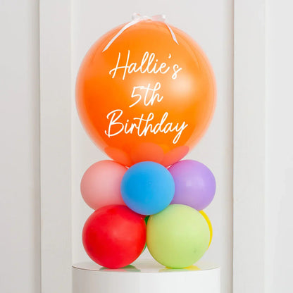 Personalised orange birthday bubble with text Hallie's 5th Birthday atop colorful rainbow balloon cluster