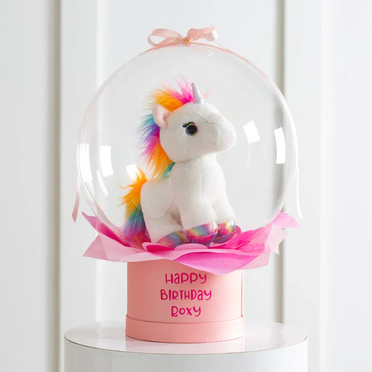 Rainbow unicorn plush toy inside clear bubble on pink box with Happy Birthday Roxy text