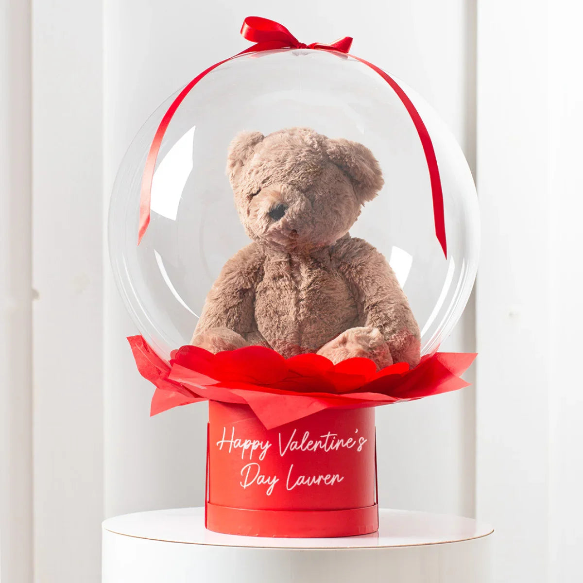 Brown teddy bear inside a clear bubble with red ribbon on red Valentine's gift box