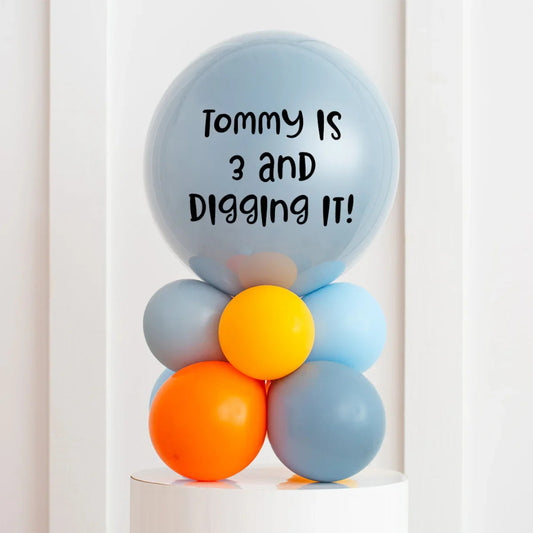 Personalised light blue bubble balloon with text 'Tommy is 3 and digging it!' atop orange, yellow, and blue balloon cluster