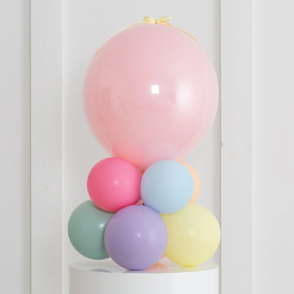 Personalised pastel rainbow air-filled bubble balloon stack on white pedestal against plain wall