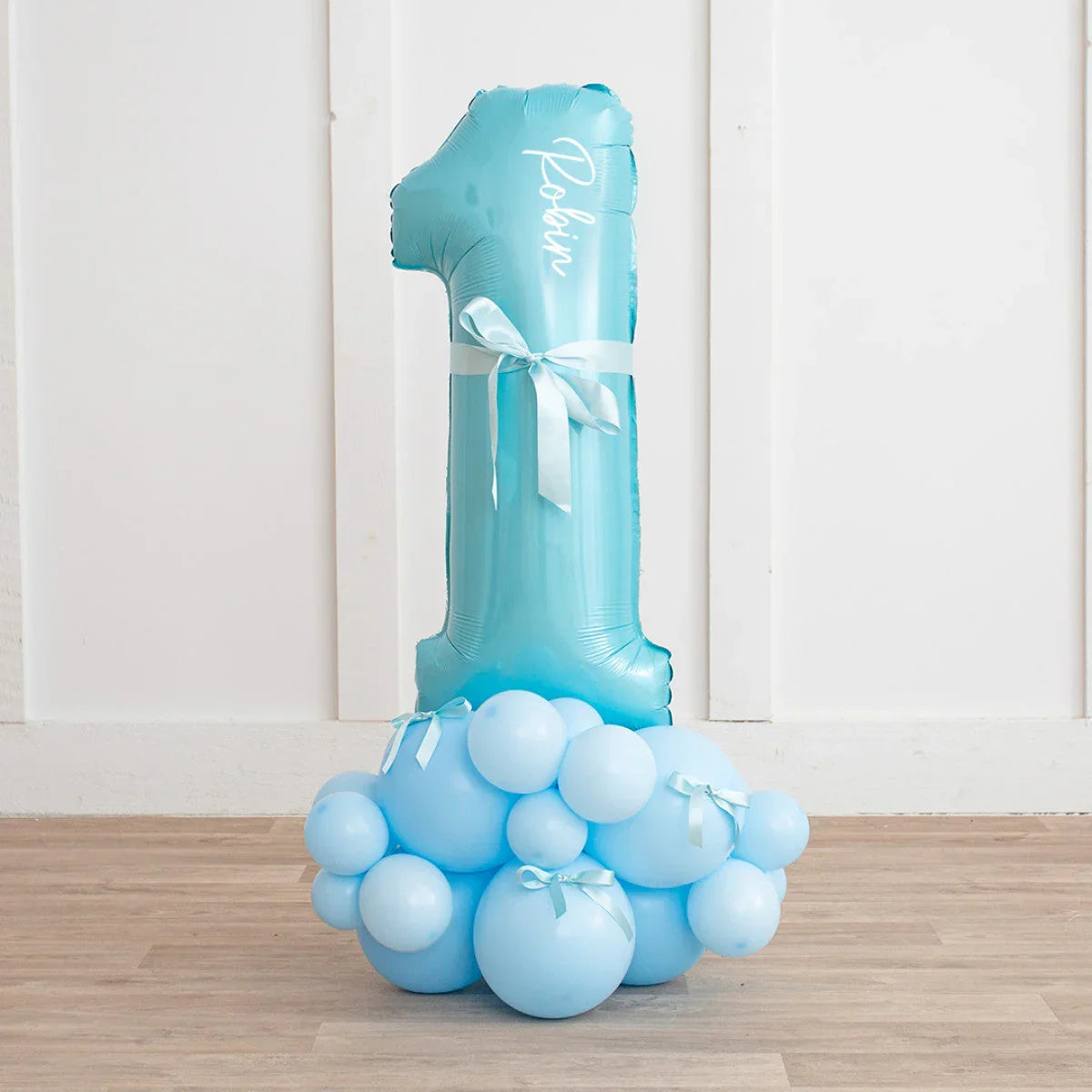 Personalized light blue number 1 balloon with ribbons atop blue balloon cluster on wooden floor