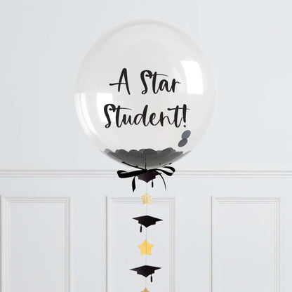 Clear graduation balloon with black confetti, ribbon, and hanging gold stars and black caps