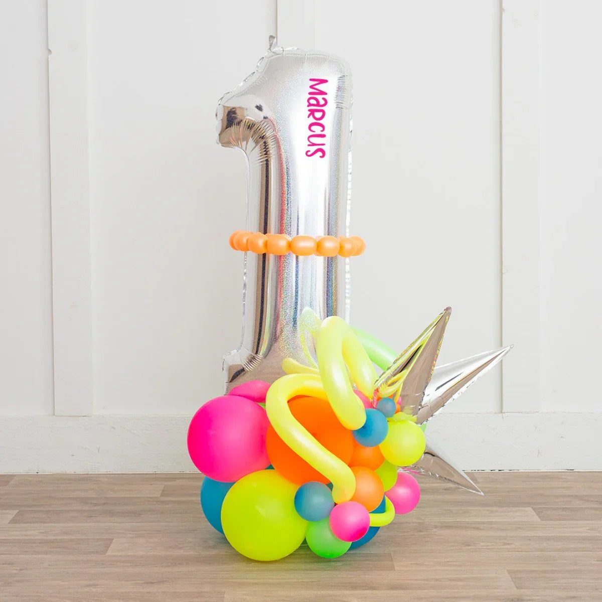 Silver number 1 balloon with personalized text Marcus surrounded by colorful neon balloons on wooden floor