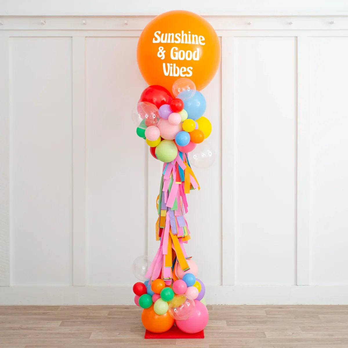 Tall colorful balloon pillar decorated with an orange balloon saying 'Sunshine & Good Vibes' indoors