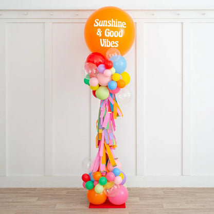 Tall colorful balloon pillar decorated with an orange balloon saying 'Sunshine & Good Vibes' indoors
