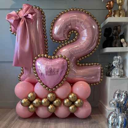 Pink and gold 12 number balloons with heart-shaped balloon and bow on pink and gold balloon base inside a room