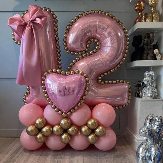 Pink and gold 12 number balloons with heart-shaped balloon and bow on pink and gold balloon base inside a room