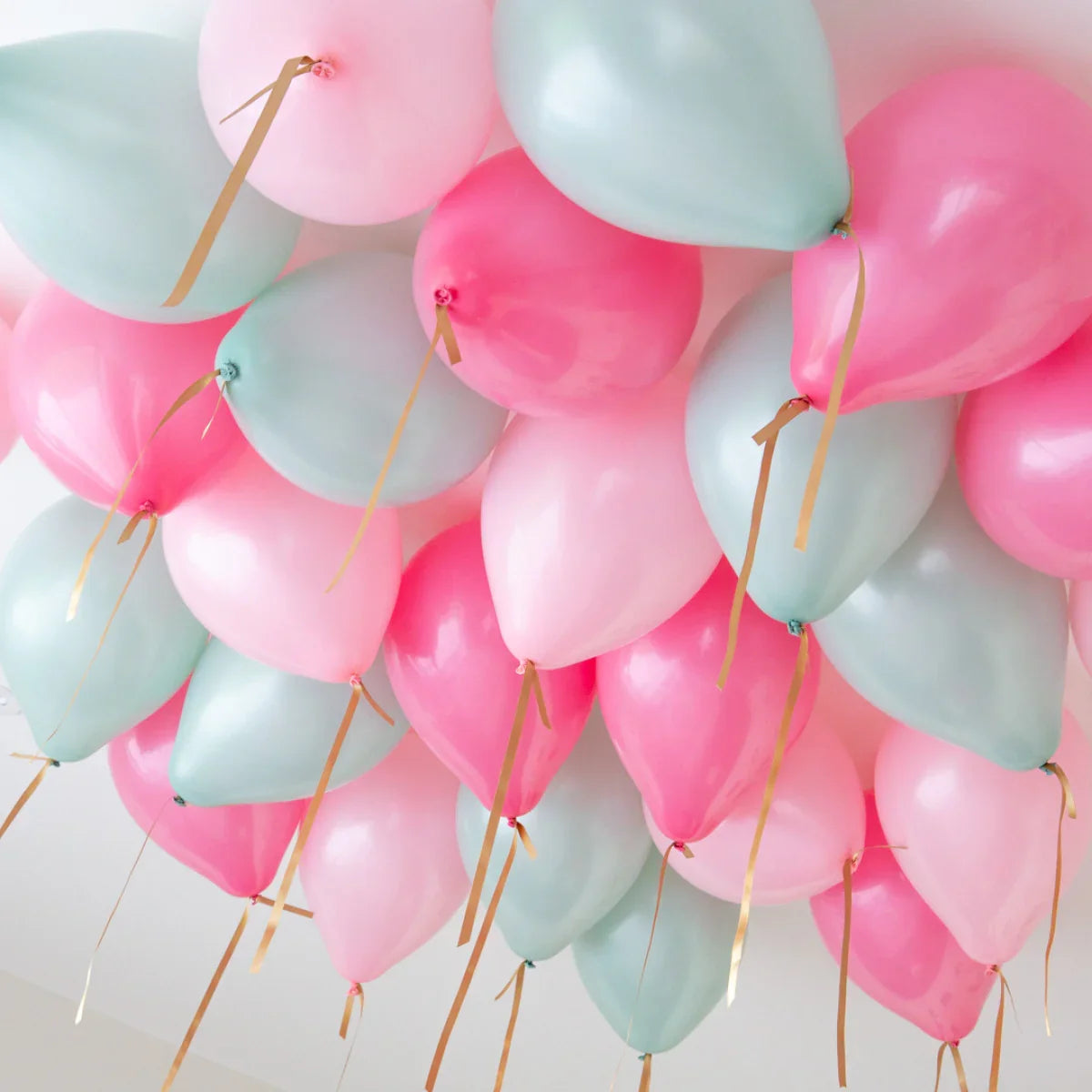 Cluster of pink, mint, and light blue helium balloons with gold ribbons floating on a white ceiling