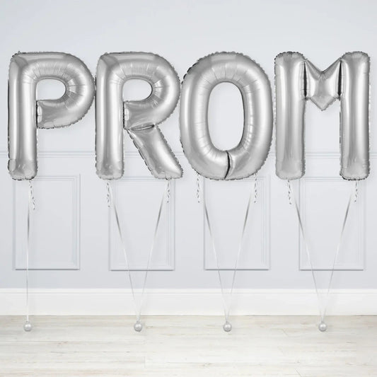 Silver foil balloons spelling PROM against a light gray wall and wooden floor