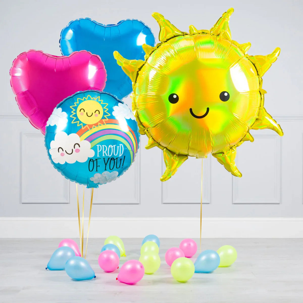 Bright helium balloons with smiling sun, heart shapes, and 'Proud of You' message in a minimalist room
