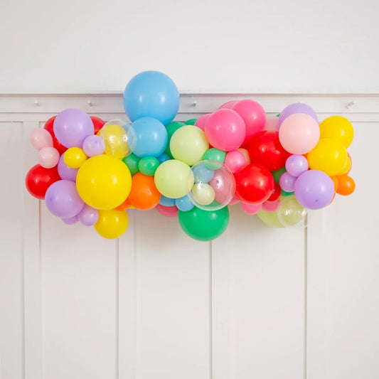 Colorful rainbow balloon garland with pink, blue, yellow, red, purple, green, and clear balloons on white wall
