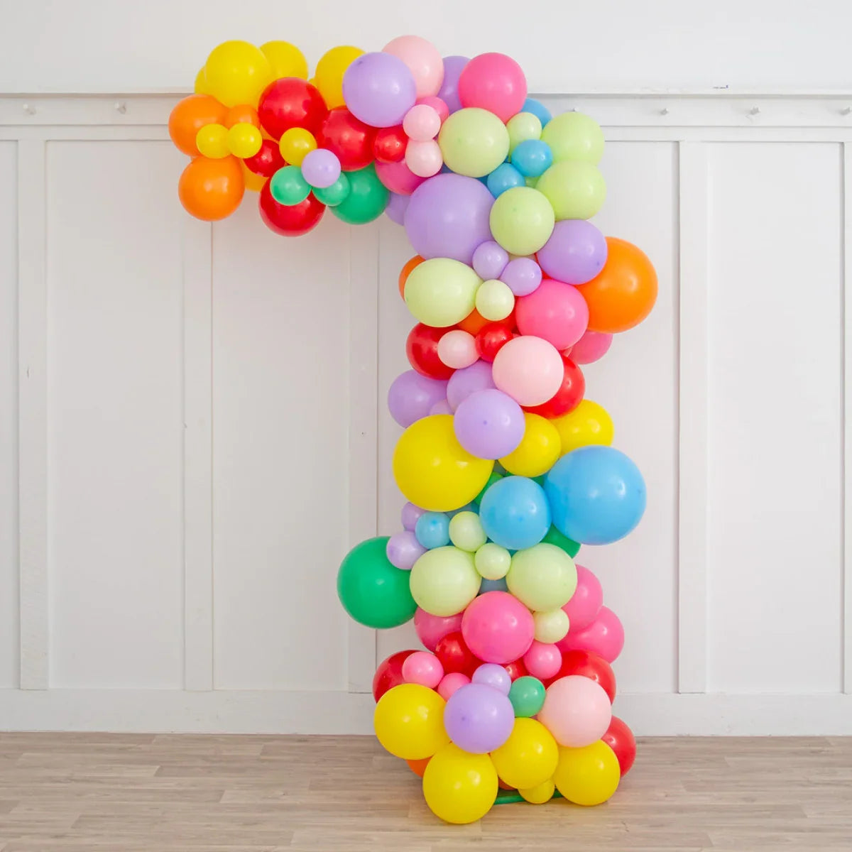 Asymmetric rainbow balloon arch decoration in pastel and bright colors against white paneled wall