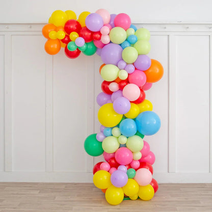 Asymmetric rainbow balloon arch decoration in pastel and bright colors against white paneled wall