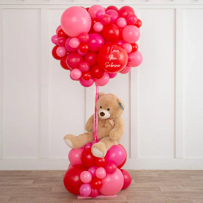 Teddy bear sitting among red and pink balloon clusters with one balloon reading I love you Sabrina