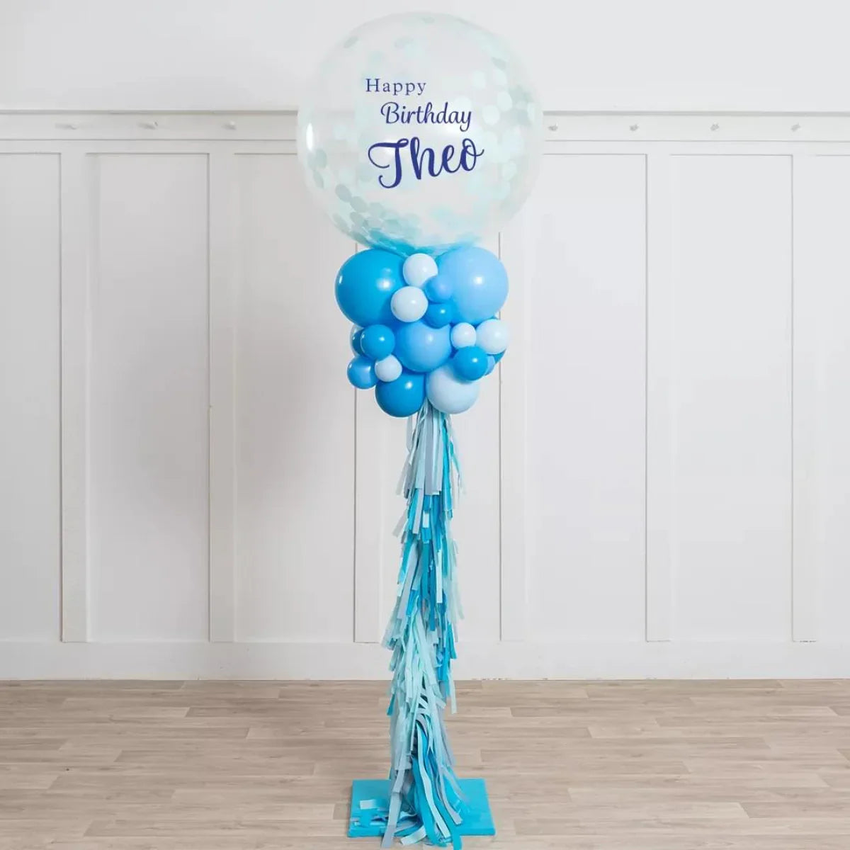 Sky blue and white birthday balloon pillar with confetti top balloon and tassels on light wooden floor