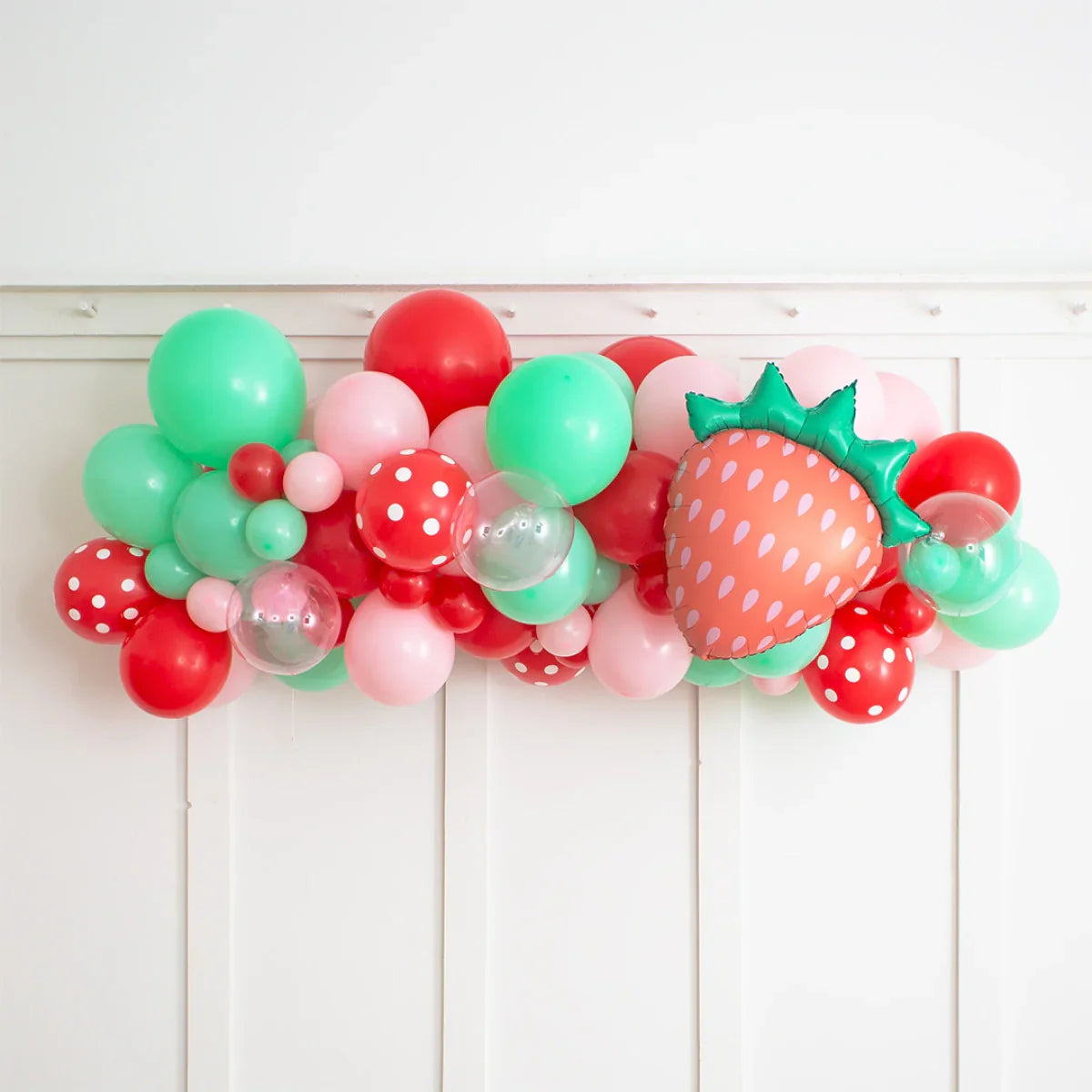 Strawberry-themed balloon garland with red, green, pink, polka dot, and transparent balloons on white wall