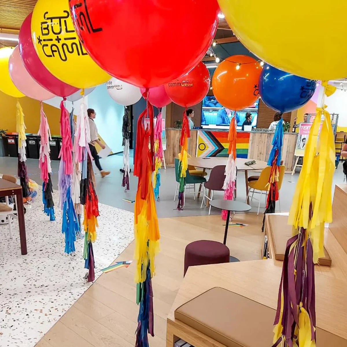 Colorful balloons with long multicolor tassels hanging in a bright, modern indoor space with seating and people