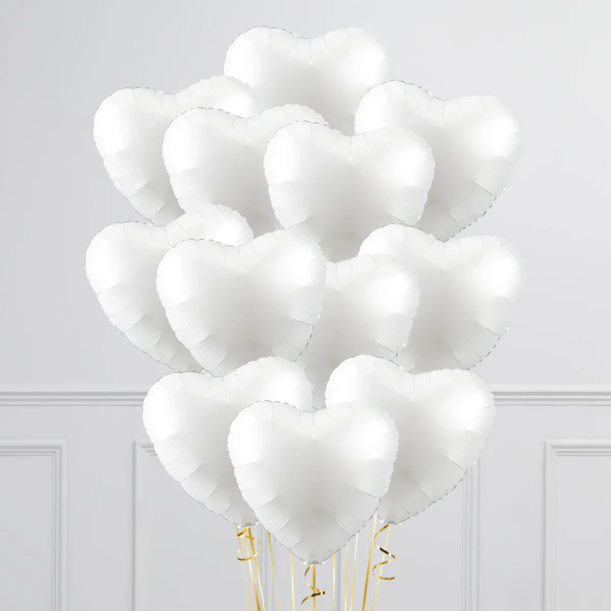 Bunch of white heart-shaped inflated foil balloons with gold ribbons against a light gray wall