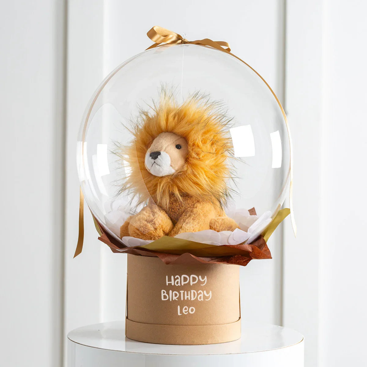 Plush lion toy inside clear balloon with golden ribbon on birthday gift box saying Happy Birthday Leo