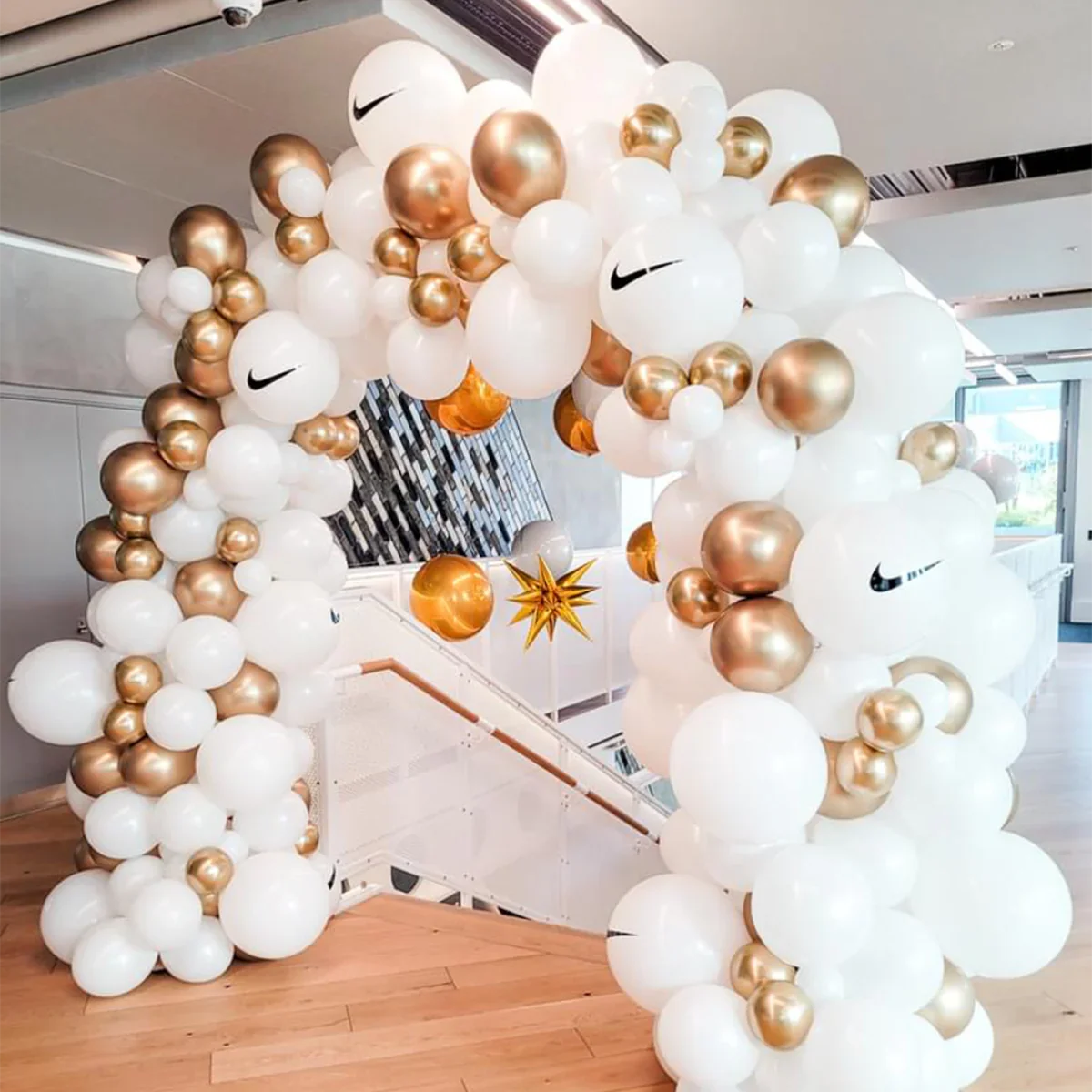 Decorative balloon arch with white and metallic gold balloons featuring Nike logos indoors near stairs