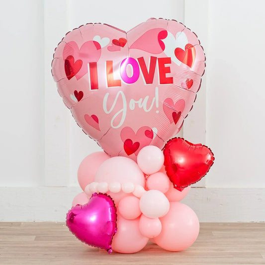 Pink heart-shaped balloon with I LOVE You text and smaller pink and red heart balloons on wooden floor