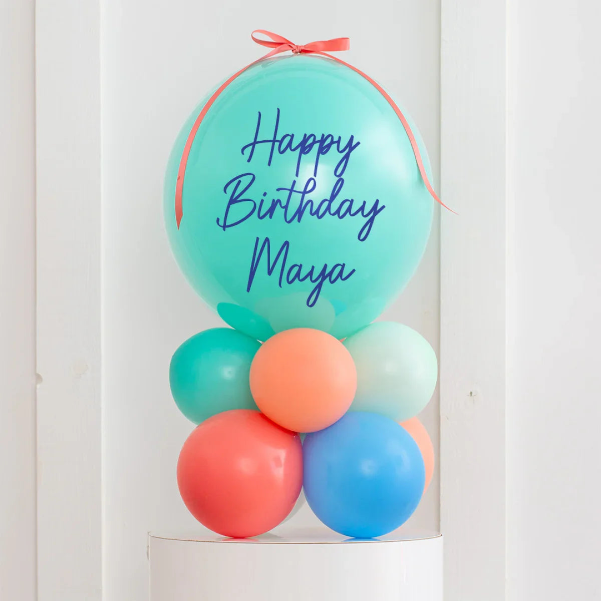 Personalized turquoise birthday balloon with blue text 'Happy Birthday Maya' and coral ribbon, surrounded by colorful smaller balloons