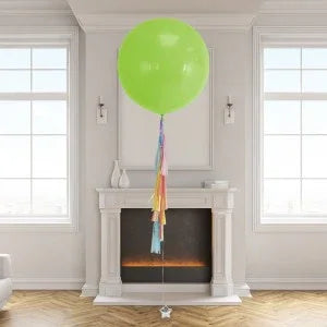 Large green helium balloon with colorful tassel tail floating indoors above fireplace in modern room