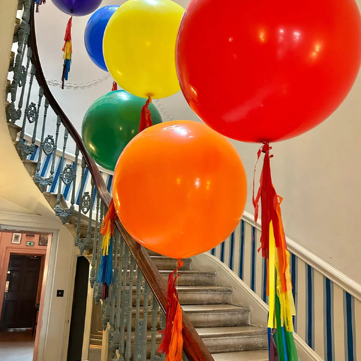 Large colorful balloons with rainbow tassels decorate a spiral staircase in a bright interior