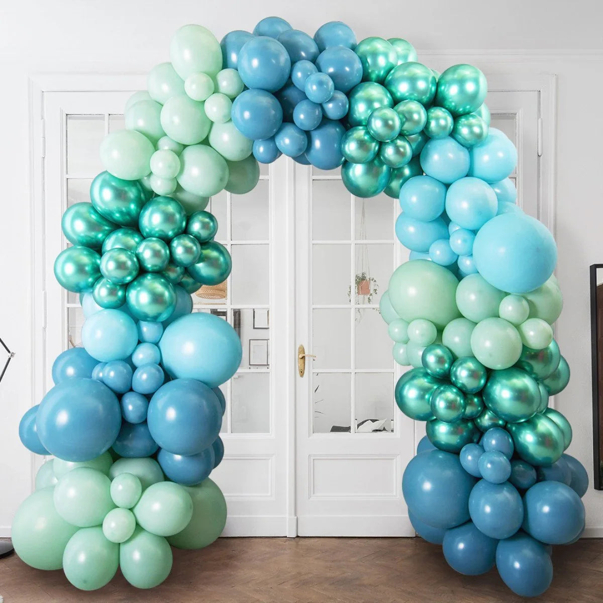 Blue and green balloon arch with matte and metallic finishes in front of white double doors indoors