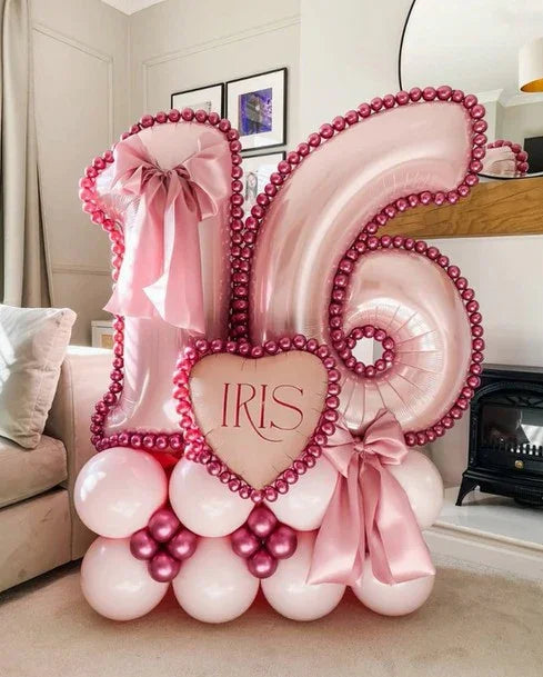 Pink metallic balloon arrangement with numbers 1 and 6, heart-shaped balloon with 'IRIS' text, and decorative bows in a living room