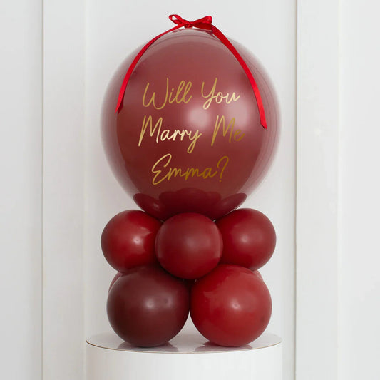Maroon marriage proposal balloon arrangement with "Will You Marry Me Emma?" text and red ribbon