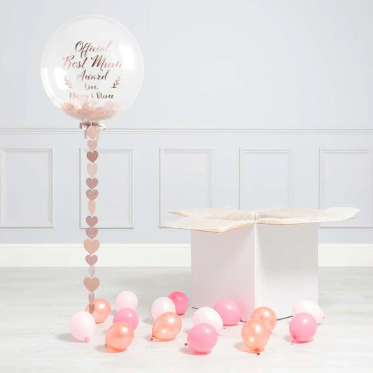 Clear balloon with Best Mum Award text, pink heart garland, pastel balloons, and white gift box