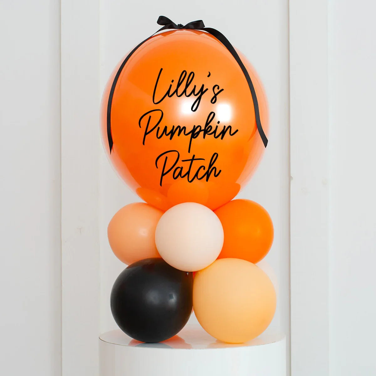 Orange balloon with black ribbon and text Lilly's Pumpkin Patch, surrounded by smaller orange, black, and beige balloons