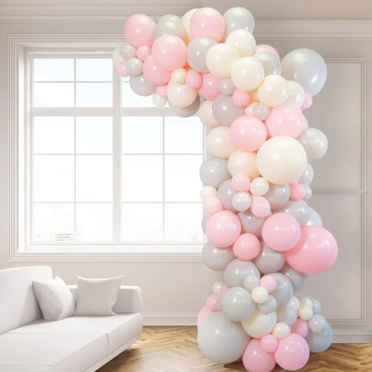 Asymmetric balloon arch in pink, gray, and white decorating a bright living room with white sofa