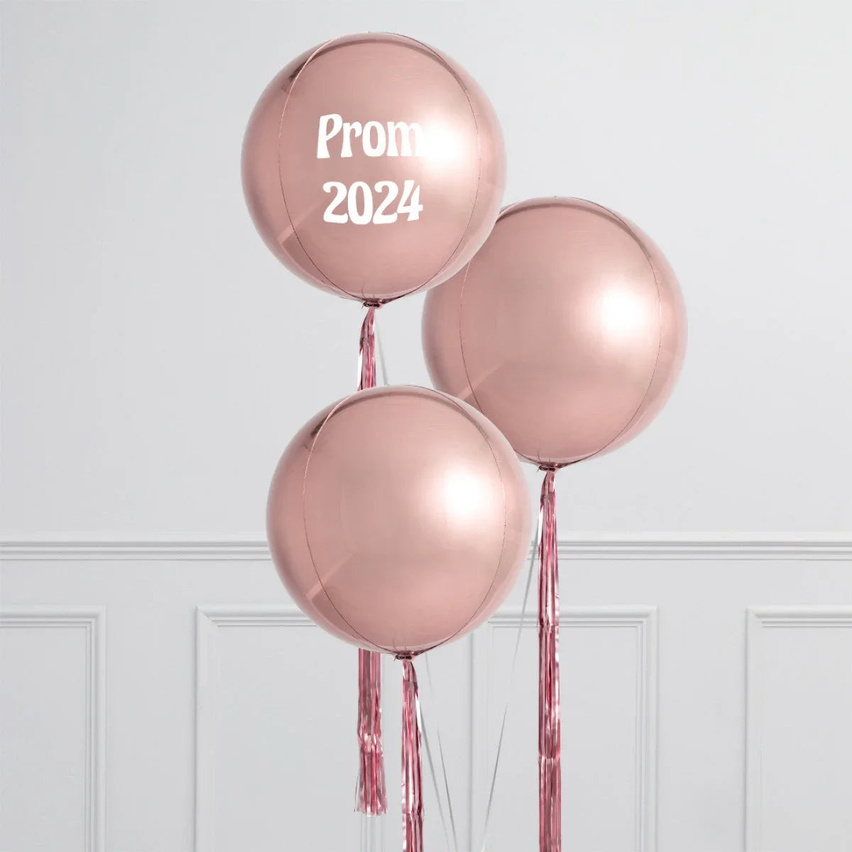 Three rose gold helium balloons with shiny tassels, one labeled Prom 2024, against white paneled wall