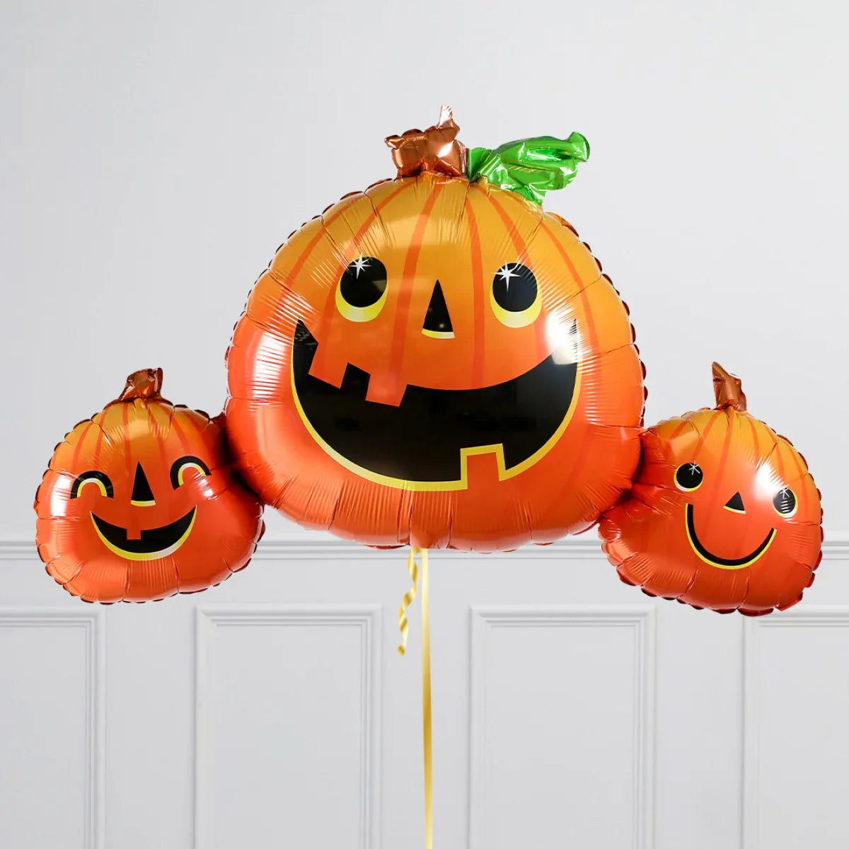 Three orange Halloween pumpkin foil balloons with smiling jack-o'-lantern faces against white wall