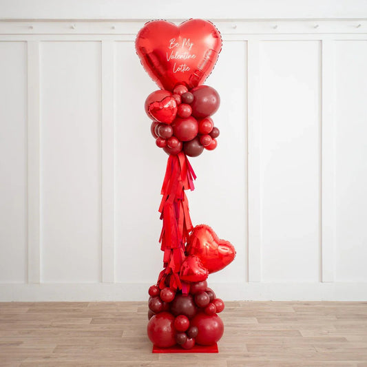 Tall red heart-shaped balloon pillar with varied balloons and red tassels in white room