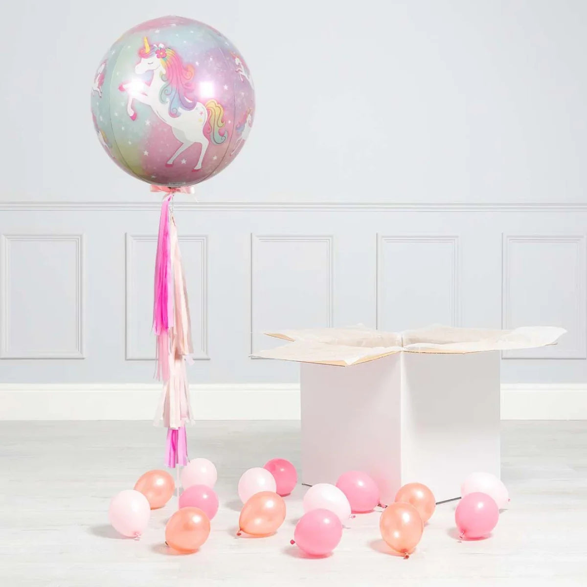 Pastel unicorn print balloon with pink tassels next to a white gift box and scattered small pink balloons
